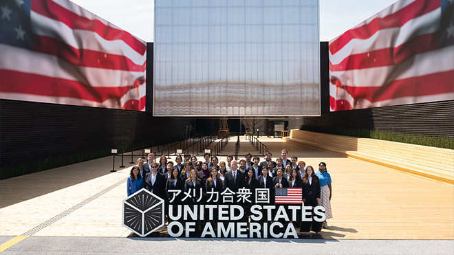 a group of people behind a sign that read "United States of America" and standing in front of a large square building with large USA flag panels on either side