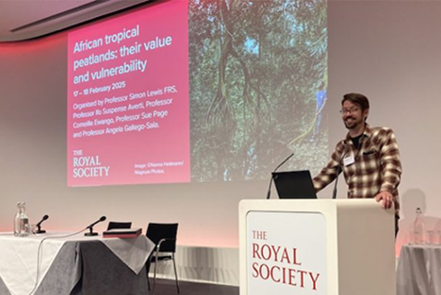 Chris Kiahtipes stands at a podium with the word Royal Society printed on it, preparing for a presentation which is projected behind him.